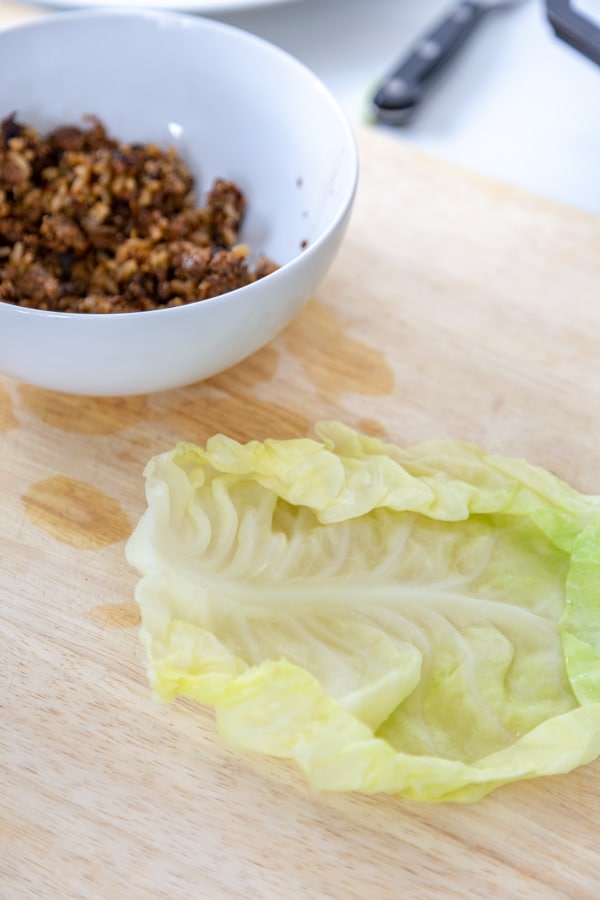 A cabbage leaf laying flat on a wood board with a white bowl of meat filling next to it.