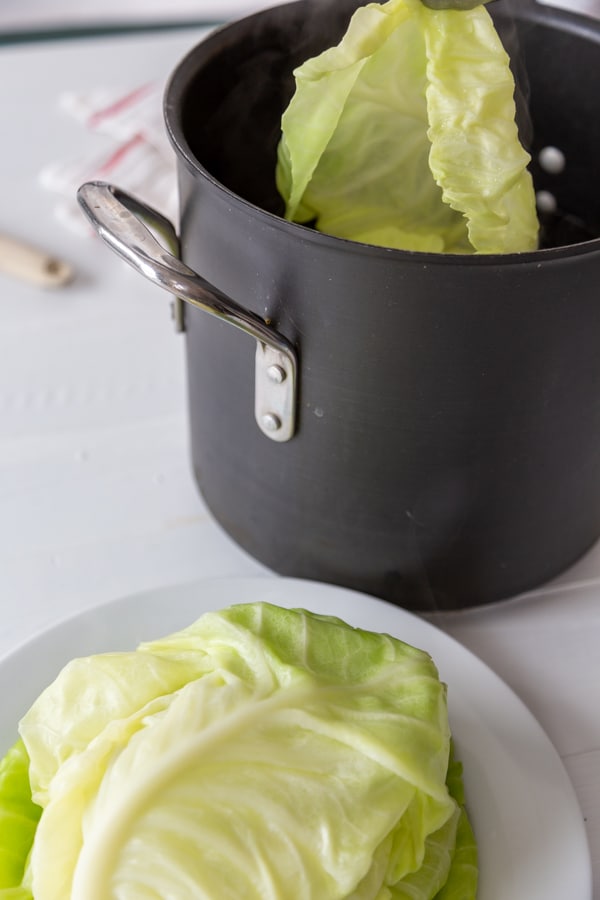 Silver tongs removing a cabbage leaf from a pot of water and a white plate with cabbage leaves is next to the pot.
