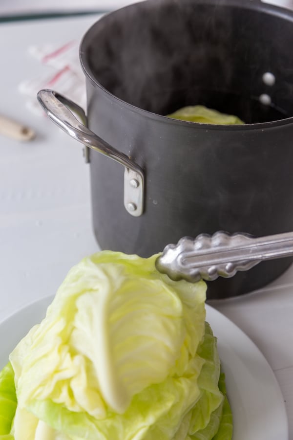 Silver tongs putting a cabbage leaf on a white plate piled with cabbage leaves.