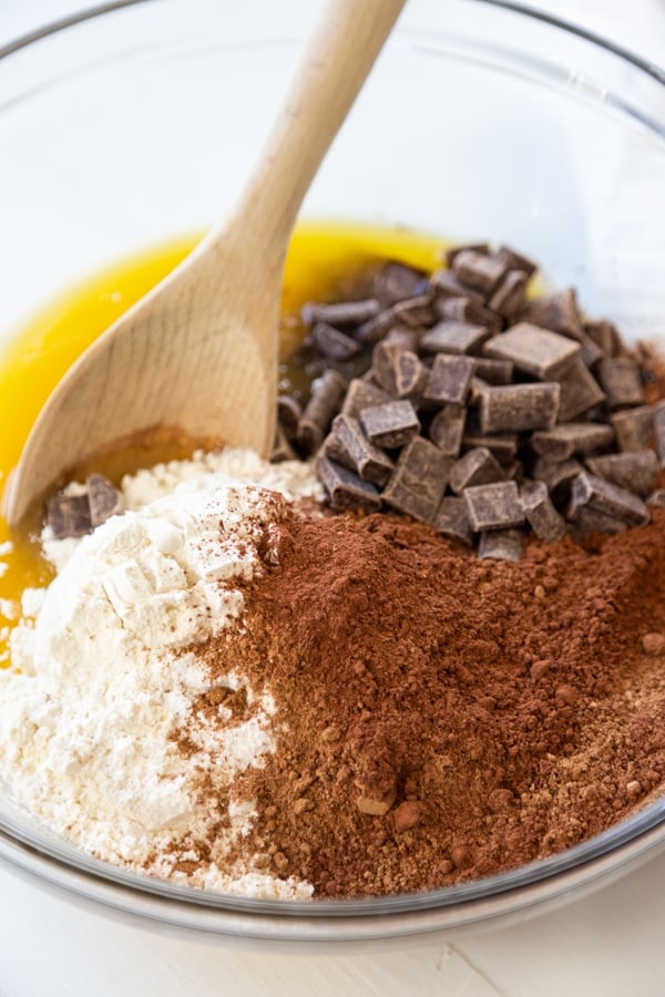 A glass mixing bowl with flour, cocoa powder, melted butter, and chocolate chunks and a wooden spoon.