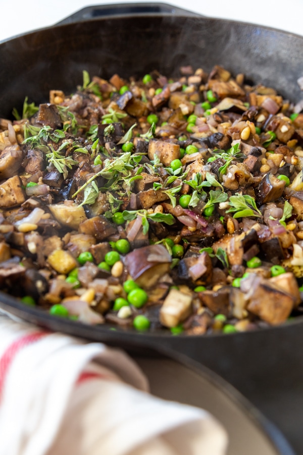 An iron skillet with eggplant, peas, onions, and pine nuts.