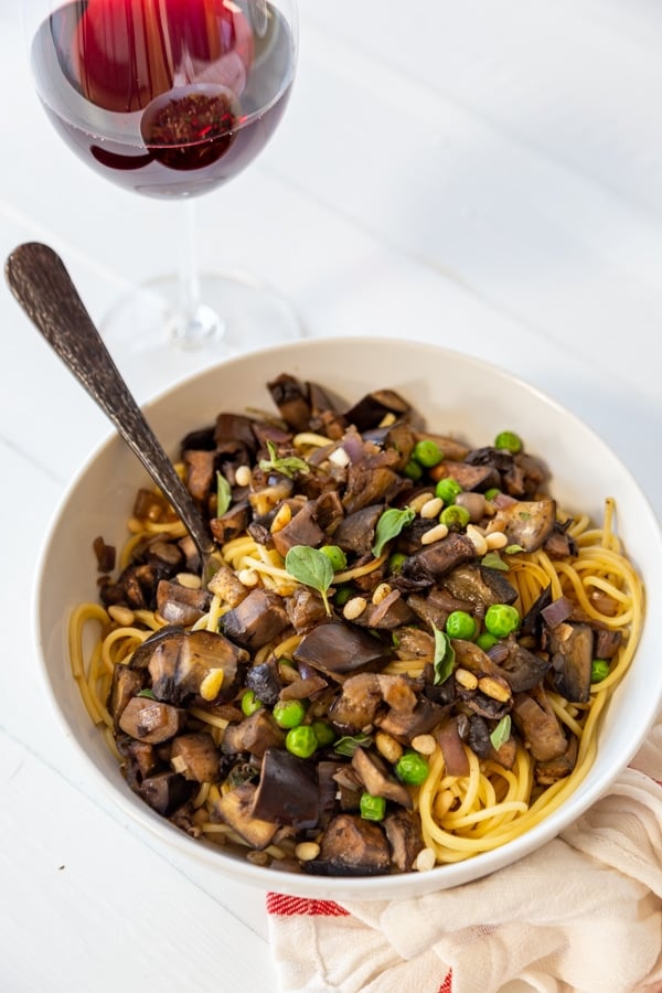 A white bowl filled with spaghetti, eggplant, peas, and pine nuts with a glass of wine behind it.