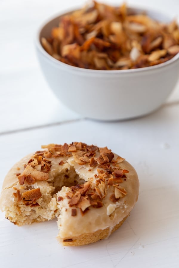 A glazed doughnut with coconut bacon on top with a bite out of it and a white bowl of coconut bacon behind it. 