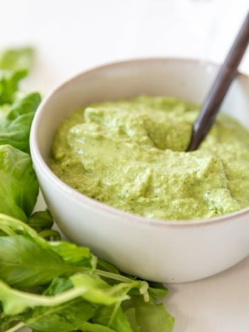 A white bowl of pesto with a spoon in the bowl and basil leaves on the left side of the bowl.