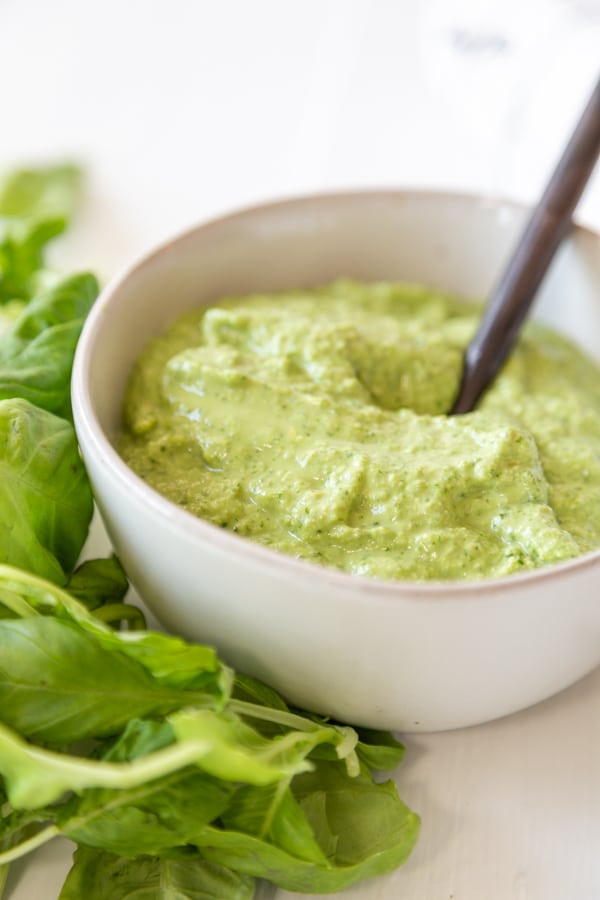 A white bowl of pesto with a spoon in the bowl and basil leaves on the left side of the bowl. 