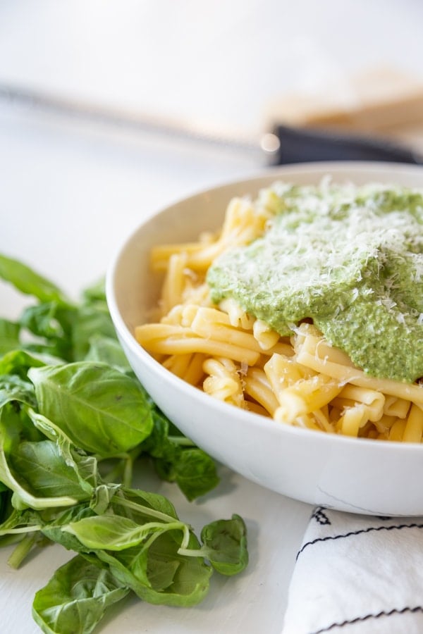 A white bowl with pasta and topped with pesto and basil leaves next to the bowl. 