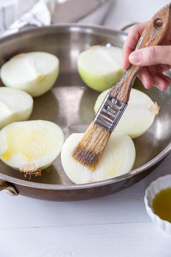 A hand brushing olive oil on halved onions. 