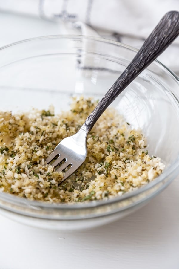 A clear glass bowl with a breadcrumb and spice mix with a silver fork in the bowl.