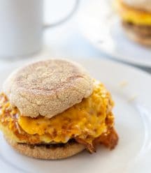 An egg and sausage breakfast sandwich on a white plate with a white coffee cup behind the plate.