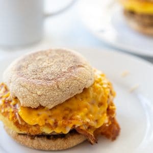 An egg and sausage breakfast sandwich on a white plate with a white coffee cup behind the plate.