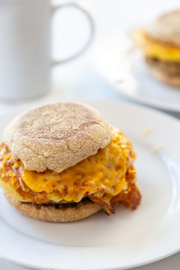 An egg and sausage breakfast sandwich on a white plate with a white coffee cup behind the plate. 