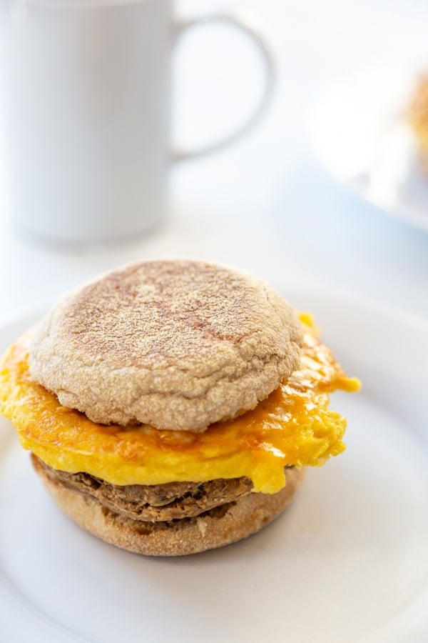 A sausage, egg, and cheese breakfast sandwich on a white plate with a white coffee cup in the background.