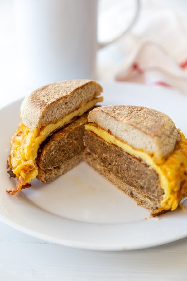 A sausage and egg breakfast sandwich cut in half on a white plate with a white coffee cup in the background. 