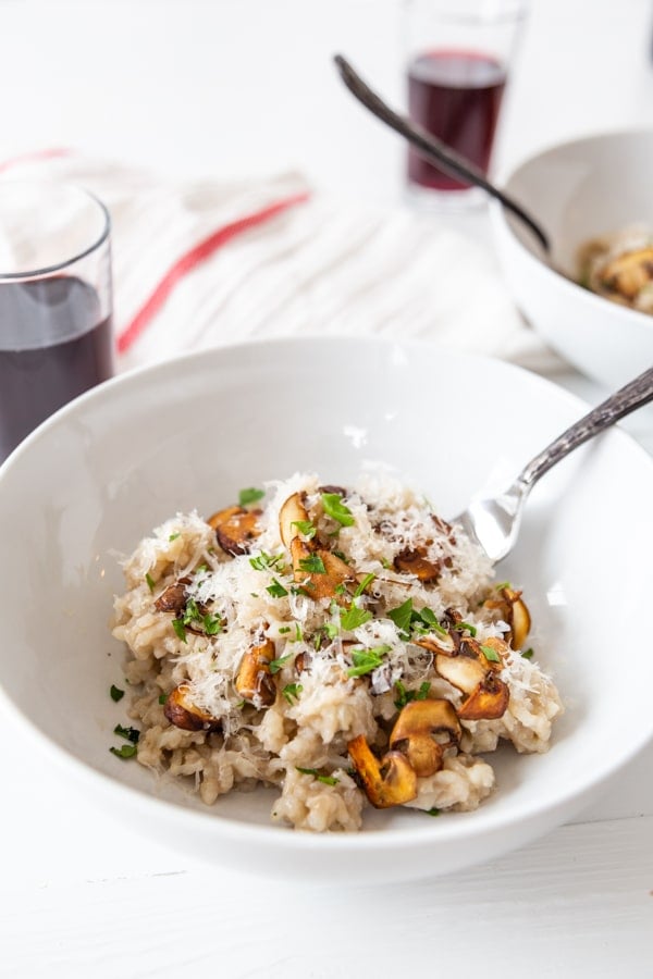 A white bowl with mushroom risotto with a fork in the bowl and a glass of wine and a white and red towel in the background.