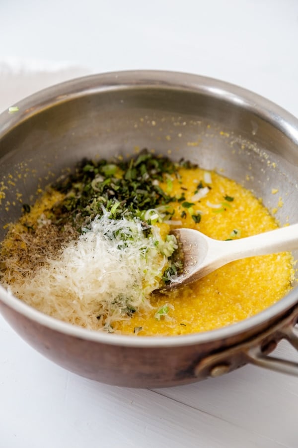 A copper saucepan with polenta and chopped herbs and grated parmesan with a wooden spoon. 