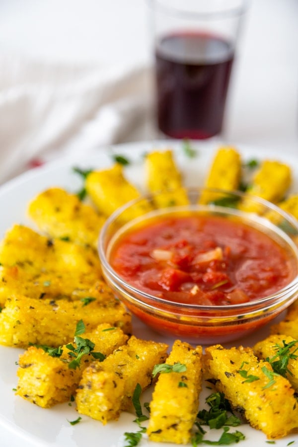 A white plate with a bowl of marinara sauce and a circle of polenta fries around it with a glass of red wine in the background. 