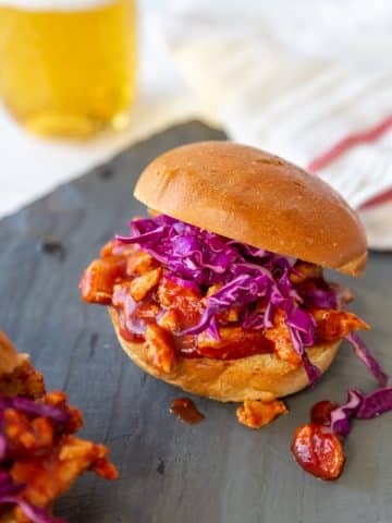 A pulled BBQ sandwich with slaw on a slate board with a beer in the background.