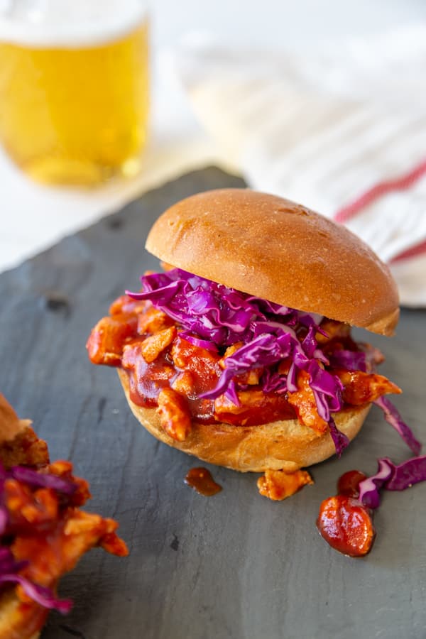 A pulled BBQ sandwich with slaw on a slate board with a beer in the background. 