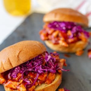 2 pulled BBQ sandwiches with purple slaw on a slate board with a glass of beer in the background.