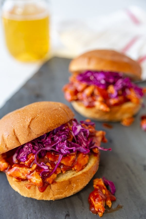2 pulled BBQ sandwiches with purple slaw on a slate board with a glass of beer in the background. 