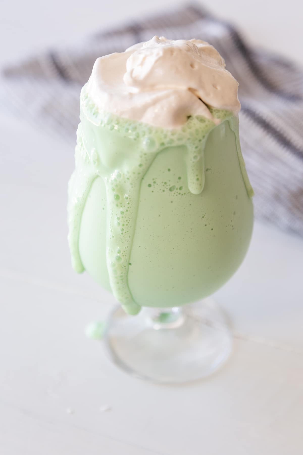 A green milkshake with the shake dripping down the side of the glass with a dollop of whipped cream on top. 