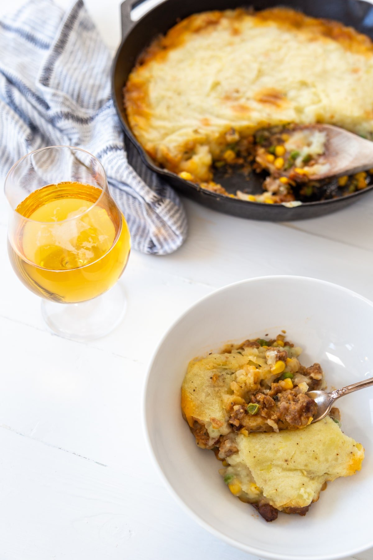 A white bowl of sheperd's pie with a fork and an iron skillet with the pie and a glass of beer in the background. 