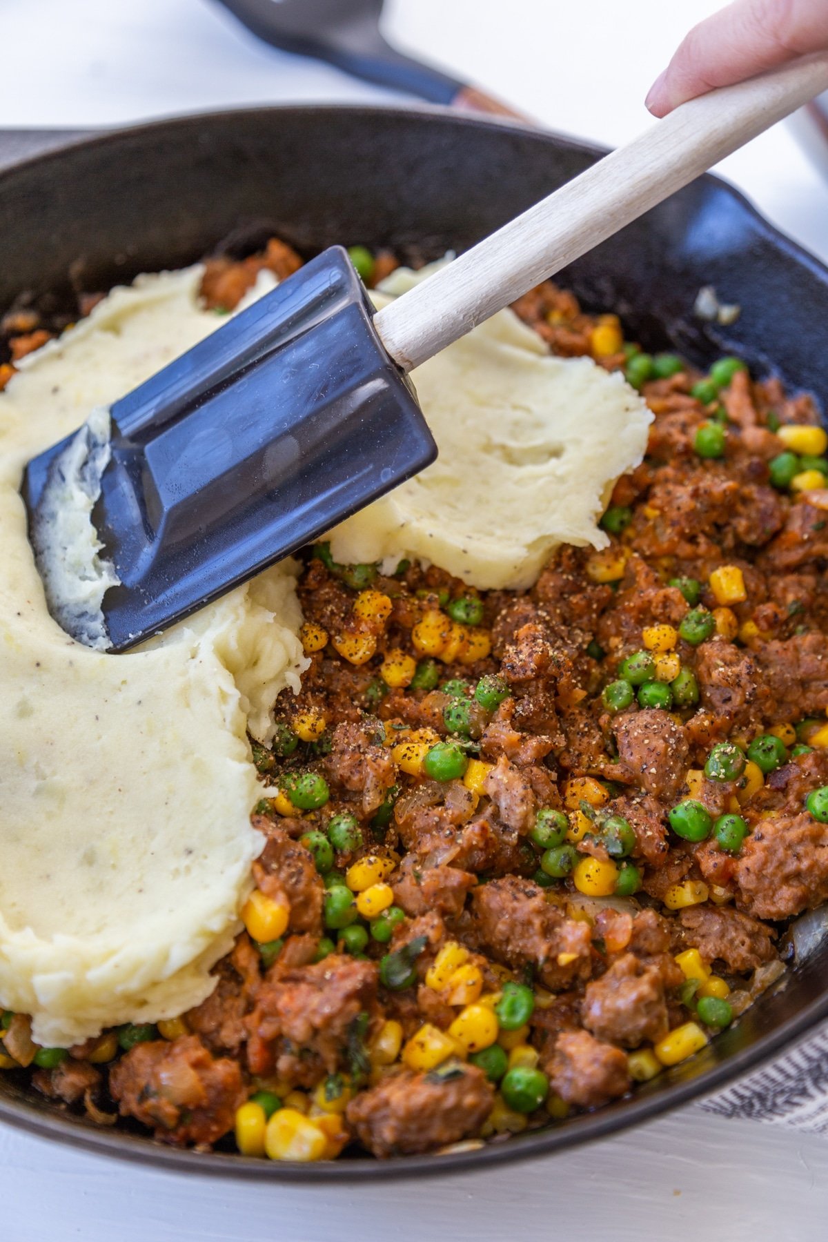 A hand with a spatula spreading whipped potatoes over a meat and vegetable filling in an iron skillet.