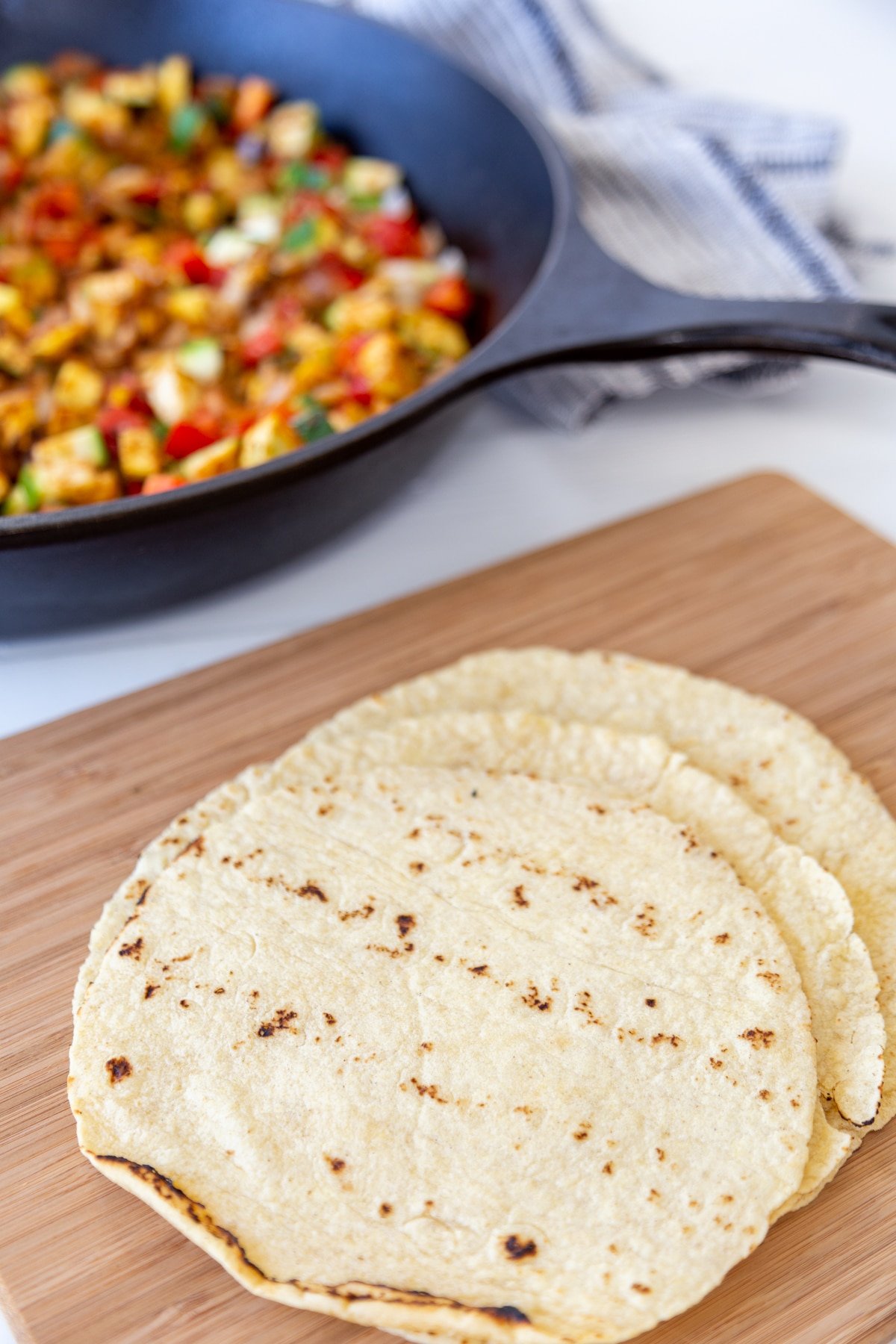 A stack of flour tortillas on a wood board with a skillet of diced veggies behind it. 