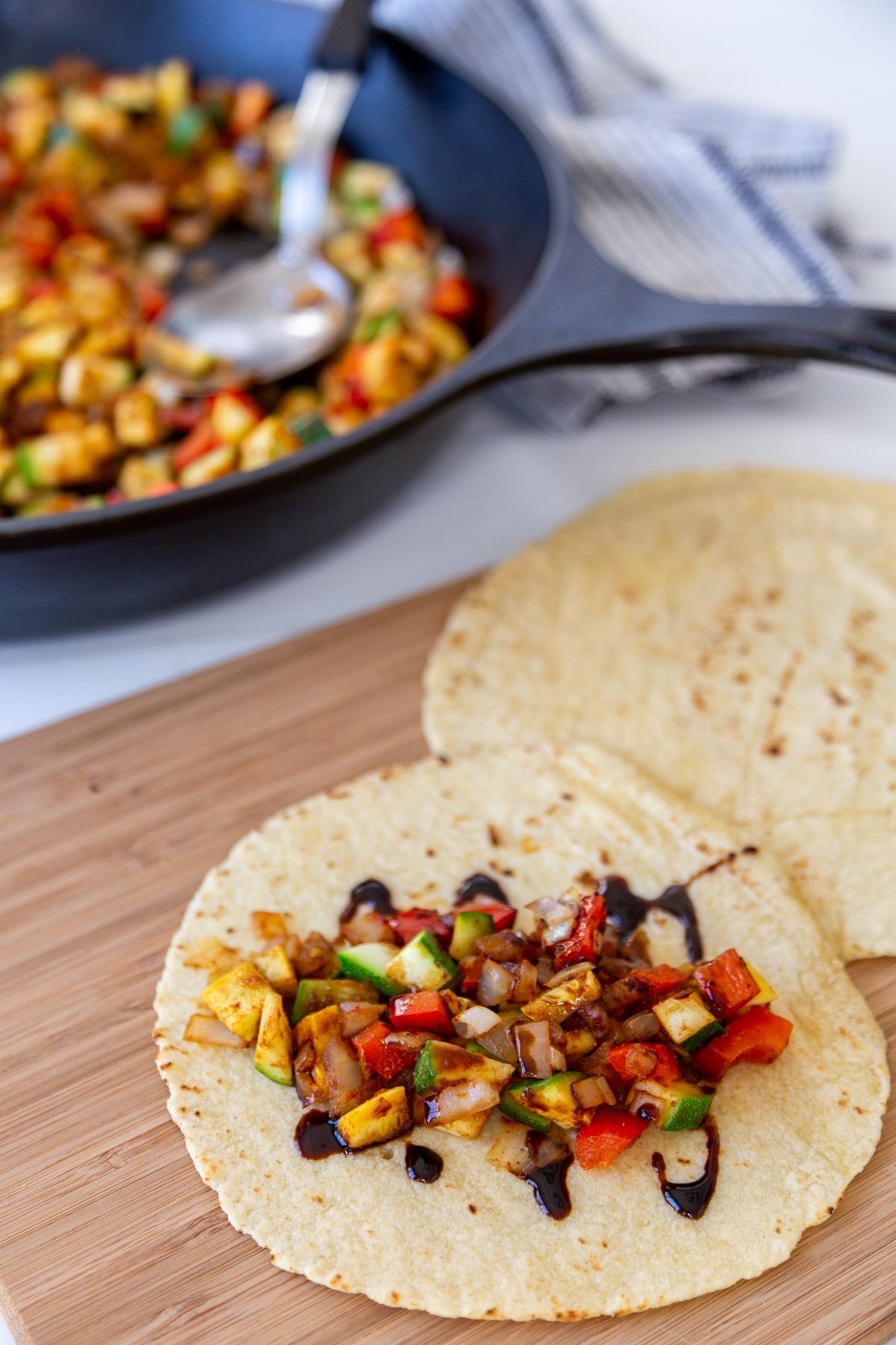 A tortilla filled with spicy veggies and drizzled with a balsamic glaze with an iron skillet with veggies behind it.