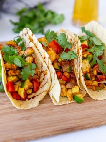 3 veggie tacos on a wood board with a glass of beer in the background.