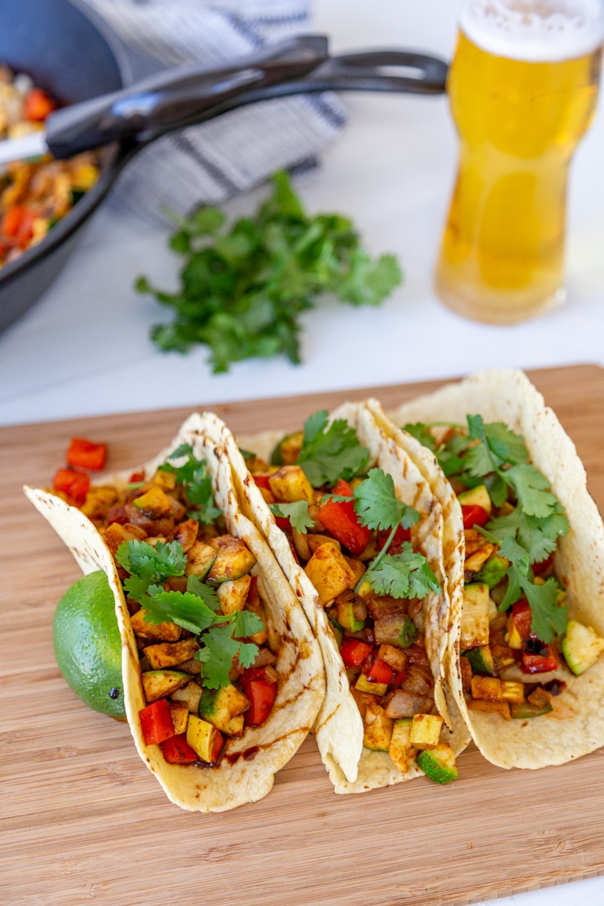 Three tacos with zucchini, squash, and bell pepper on a wood board with a beer and the skillet of veggies in the background. 