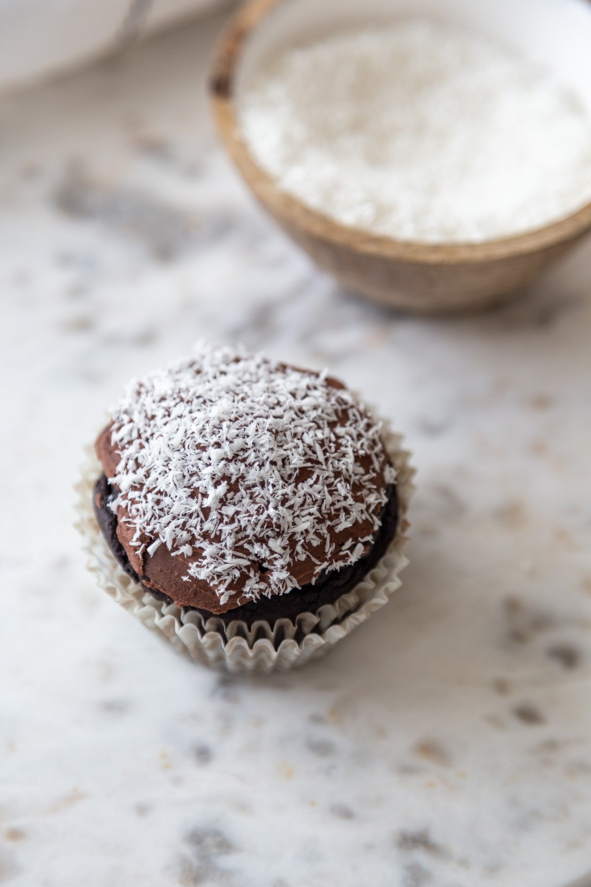 A chocolate coconut cupcake on a marble board with a bowl of shredded coconut behind it.