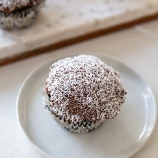 A chocolate frosted cupcake dipped in shredded coconut on a white plate, with a marble board of cupcakes in the background.