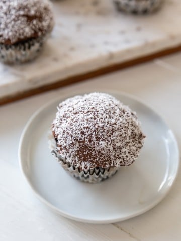 A chocolate frosted cupcake dipped in shredded coconut on a white plate, with a marble board of cupcakes in the background.