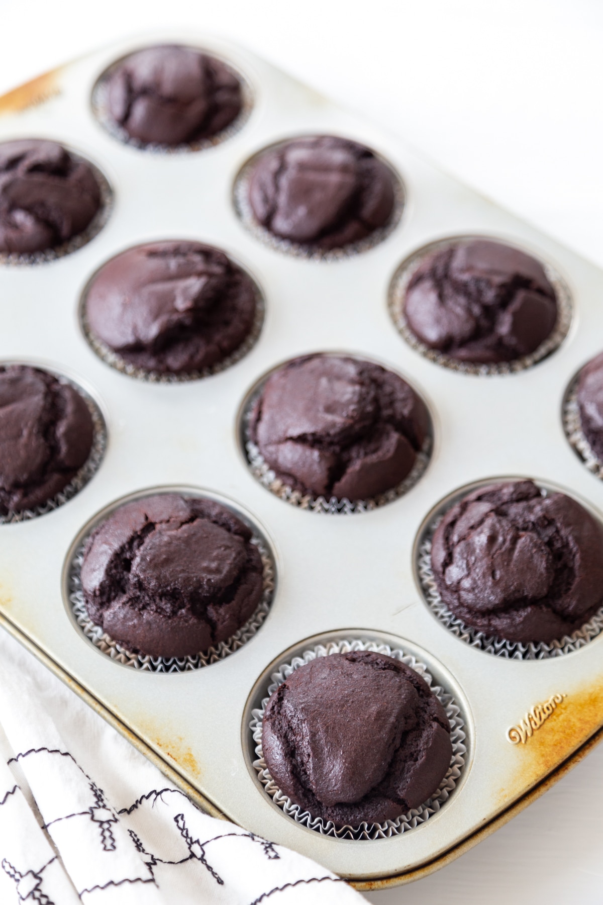 A muffin tin with chocolate cupcakes and a white and black towel next to the tin.