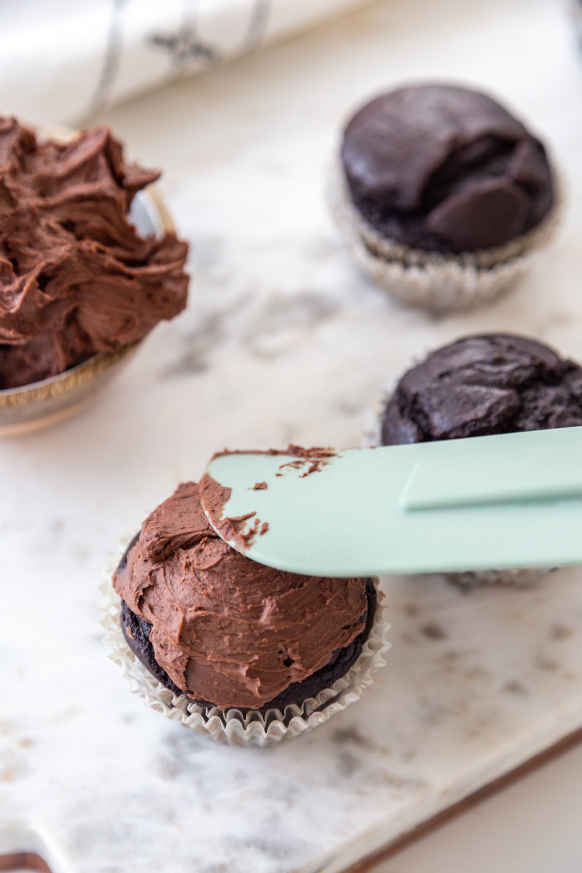 A green spatula spreading chocolate frosting on a chocolate cupcake.