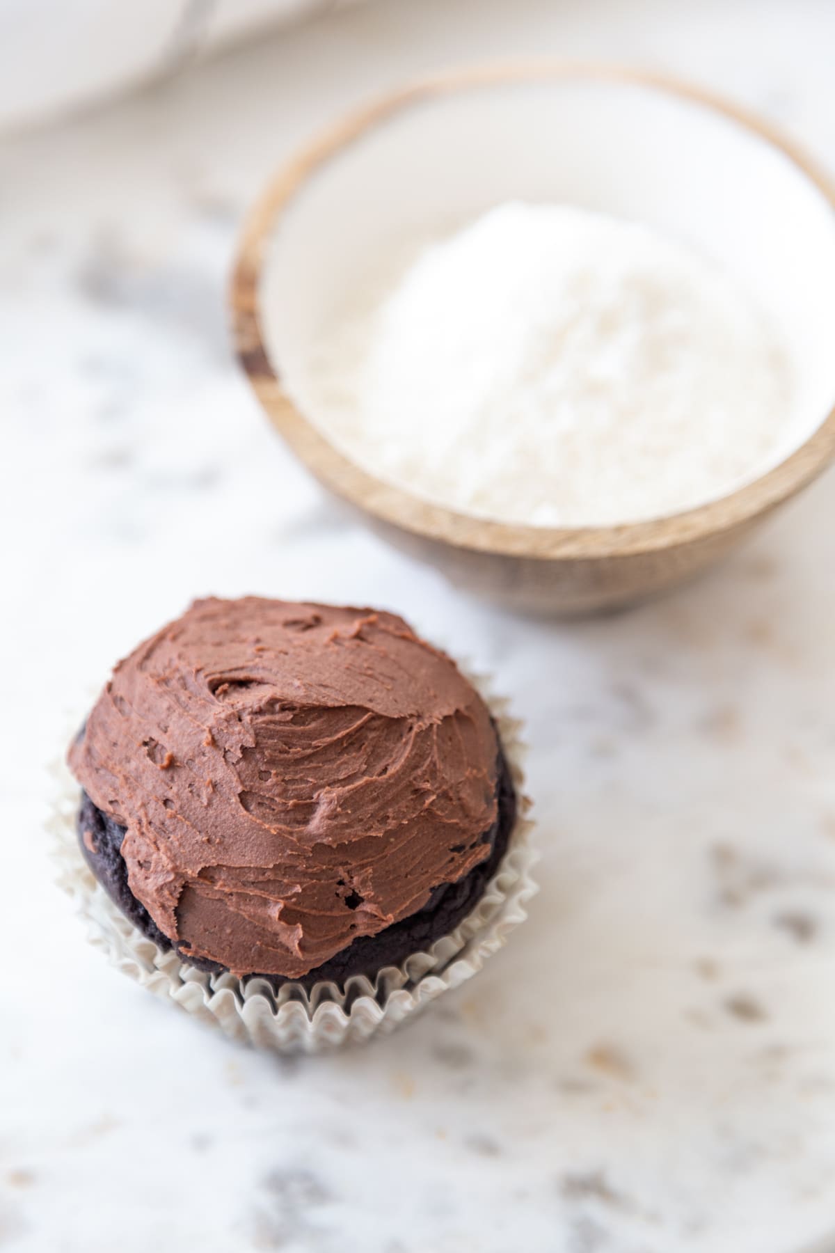 A chocolate cupcake with chocolate frosting and a bowl of shredded coconut next to the cupcake.
