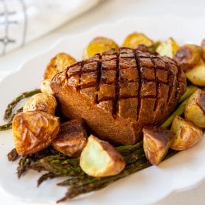 A sliced vegan ham on a bed of asparagus on a white platter with potatoes.