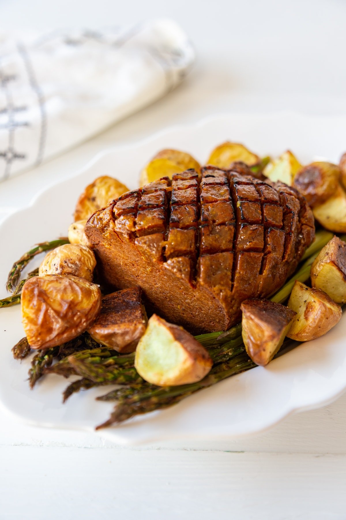 A sliced vegan ham on a bed of asparagus on a white platter with potatoes. 