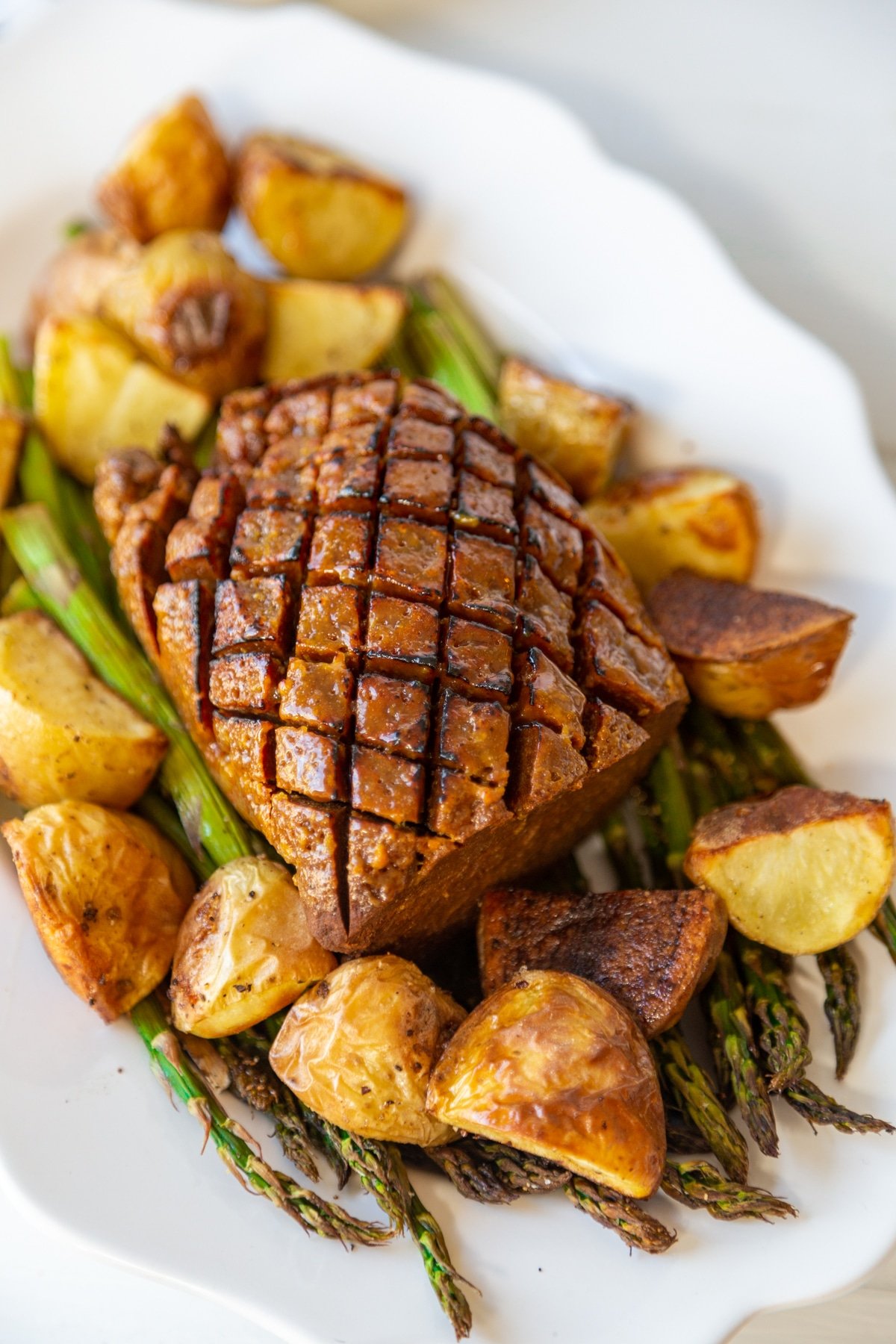 An overhead shot of a seitan ham that's scored on the top with roasted potatoes and a bed of roasted asparagus. 