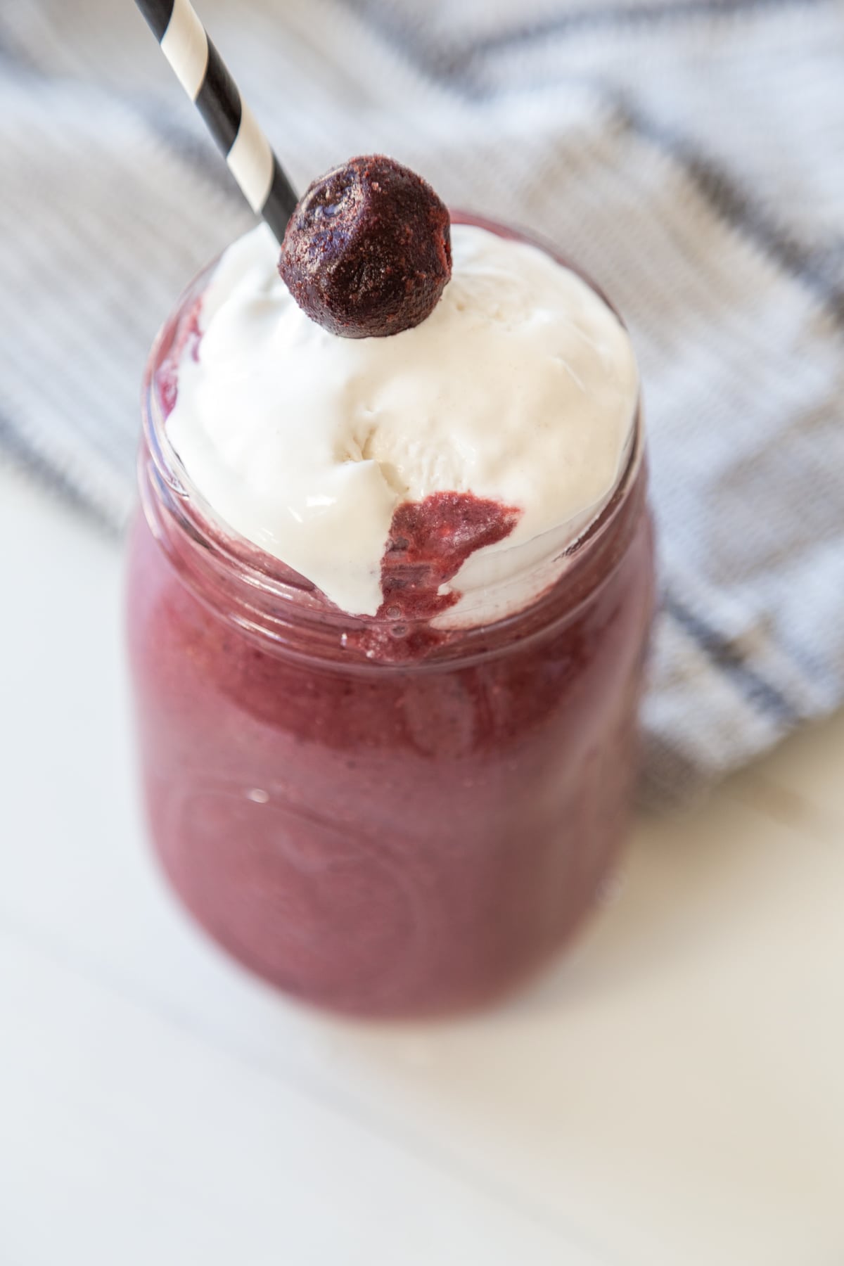 A cherry smoothie in a mason jar with whipped topping and a cherry on top, and a black and white straw in the glass. 