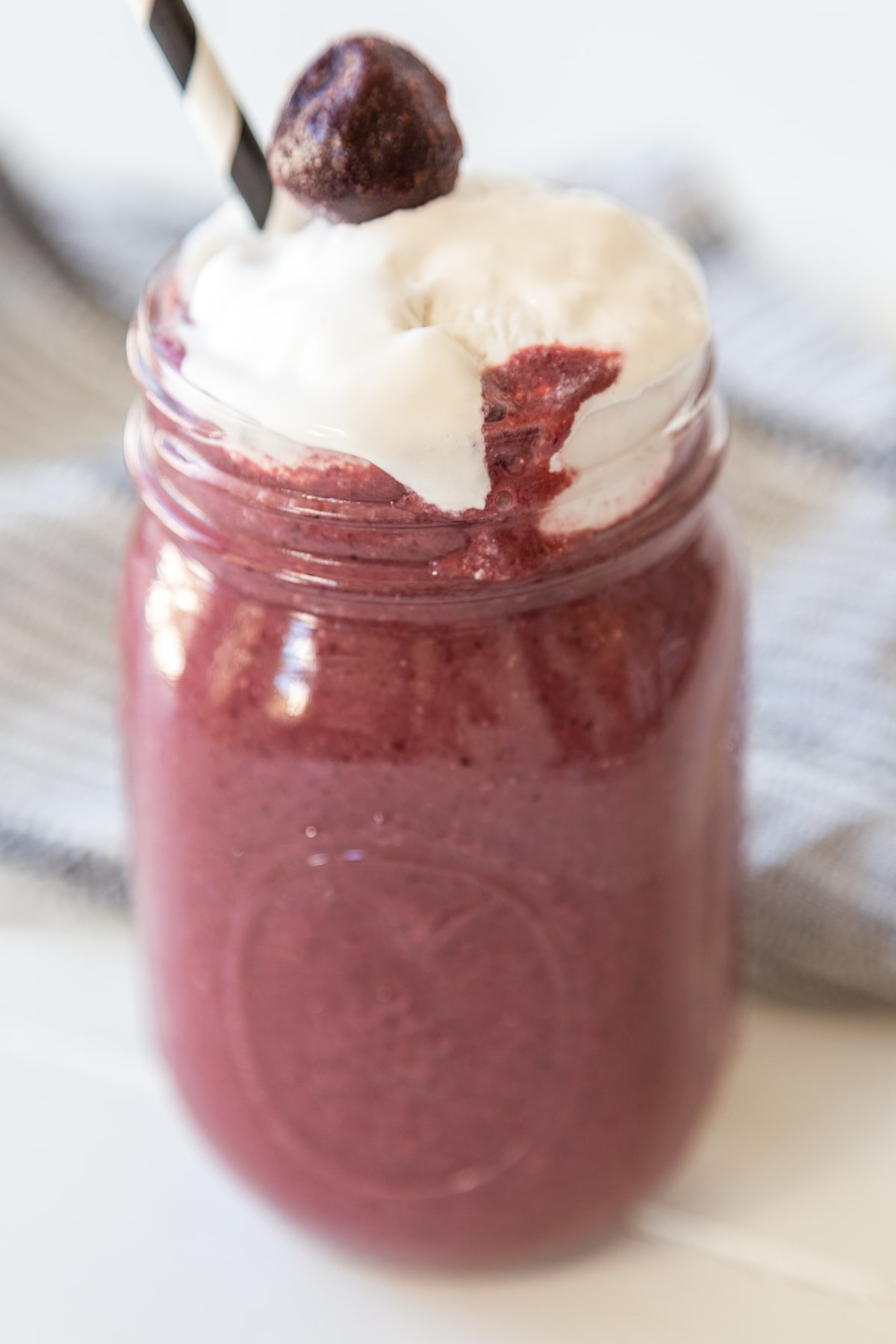 A cherry smoothie with whipped topping and a cherry on top in a mason jar with a black and white straw. 