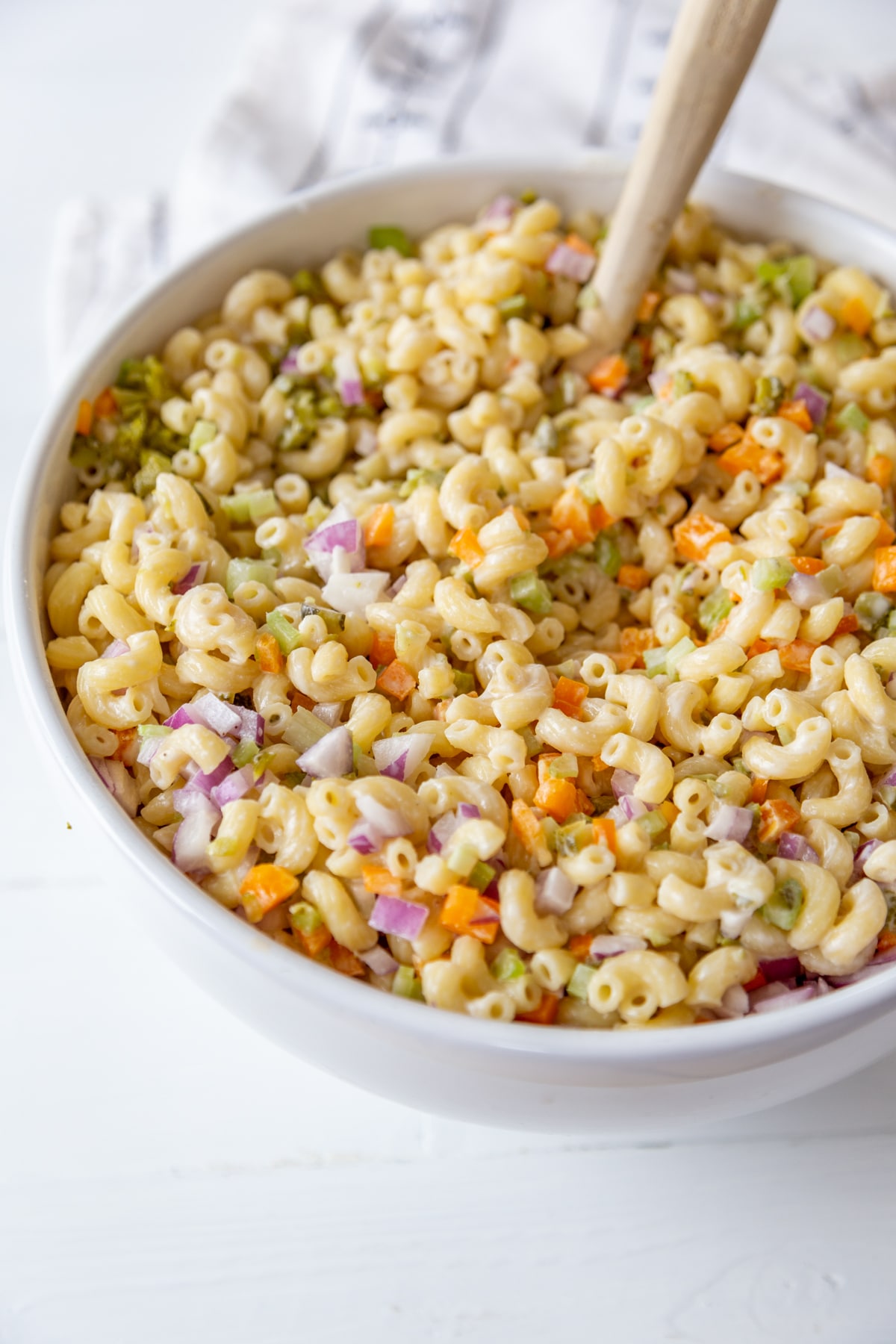 A large white bowl with a wooden spoon stirring macaroni salad.