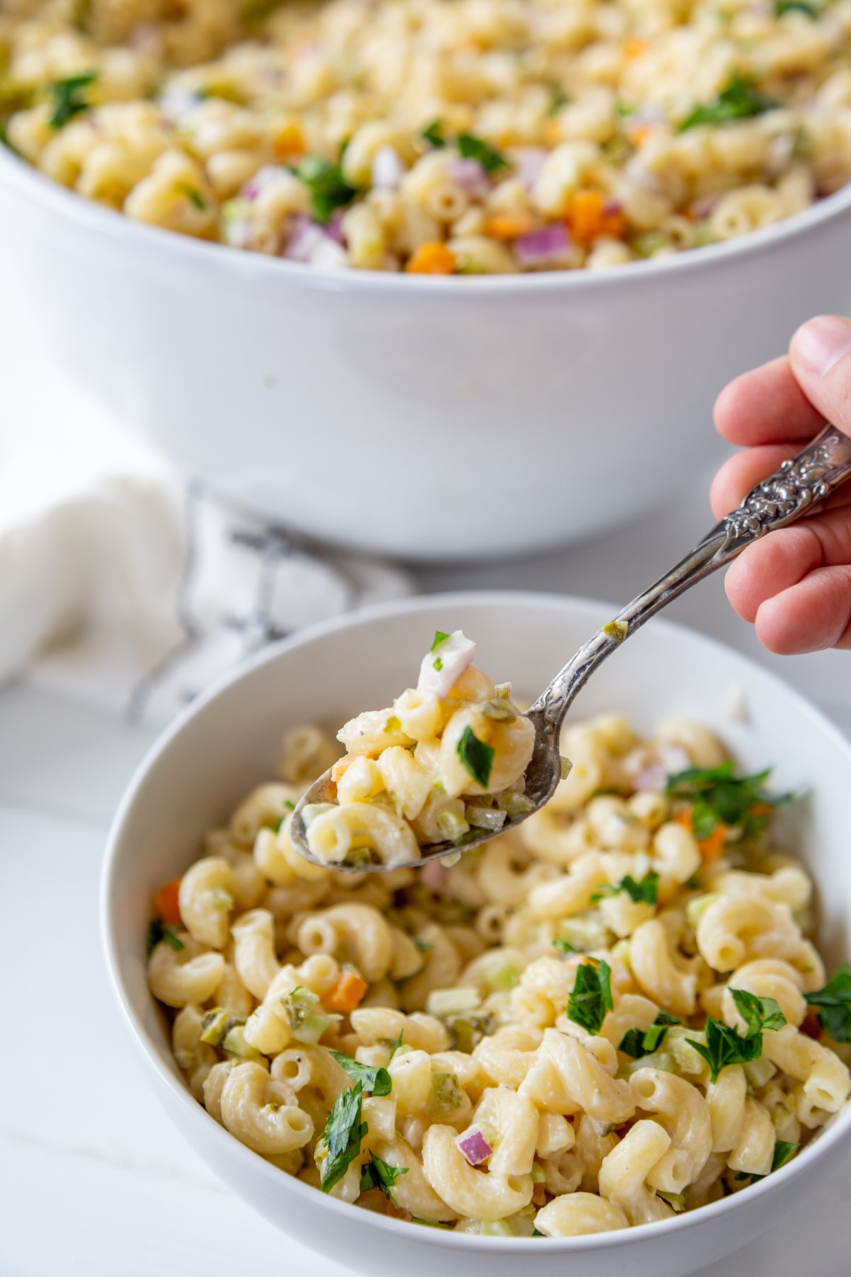 A white bowl with macaroni salad and a hand with a silver fork filled with the salad, and a large bowl of the salad in the background. 