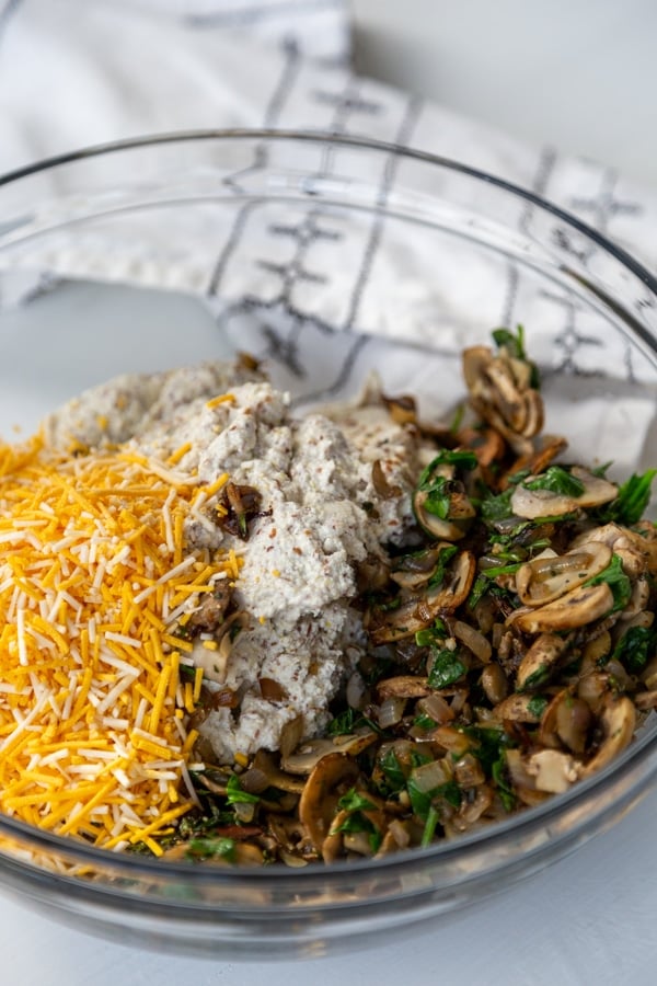 A glass mixing bowl with cheese, mushrooms, onions, spinach, and tofu.