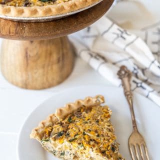 A slice of vegetable quiche on a white plate with a silver fork and a wooden cake stand with the entire quiche behind it.