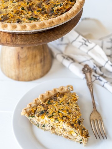 A slice of vegetable quiche on a white plate with a silver fork and a wooden cake stand with the entire quiche behind it.