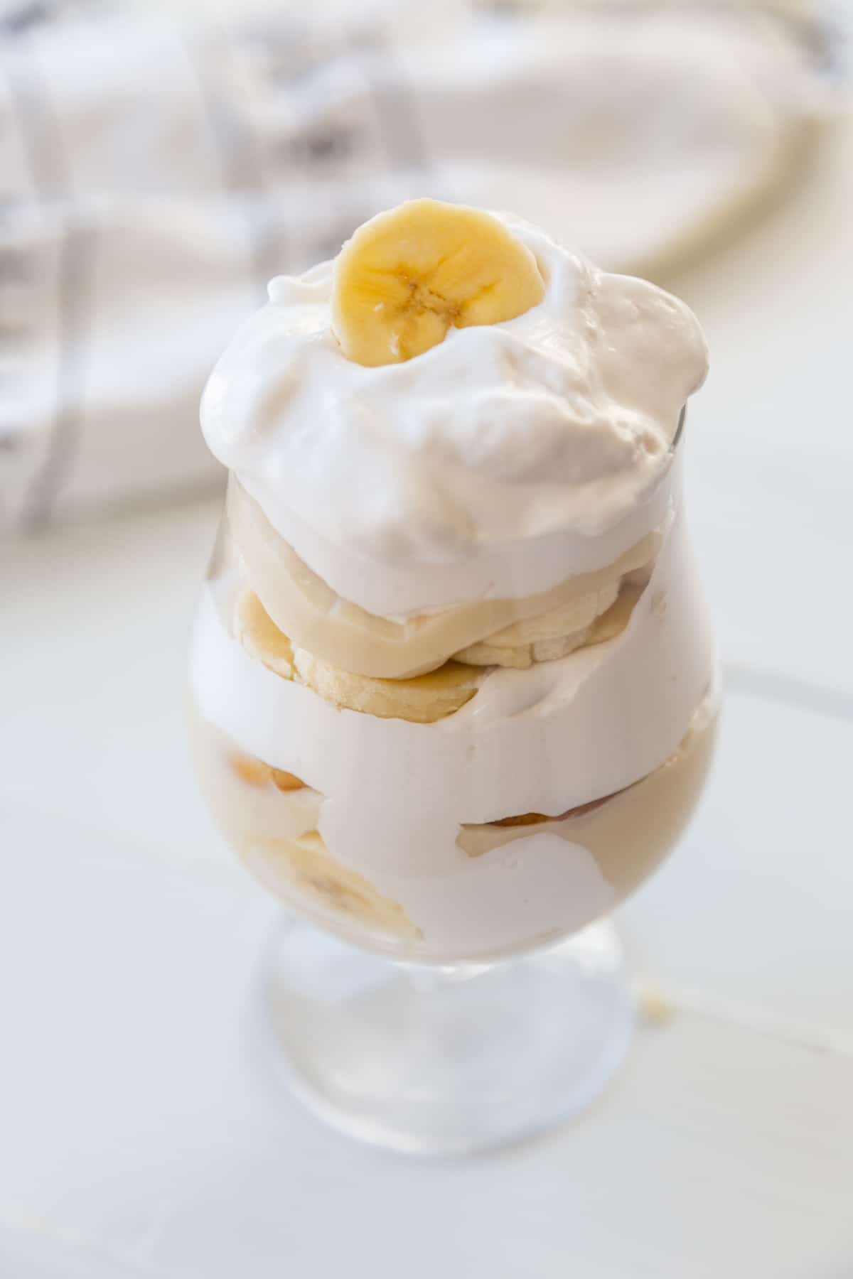 A glass filled with banana pudding, vanilla wafers, and whipped cream, with a silver spoon and a banana slice on top. 