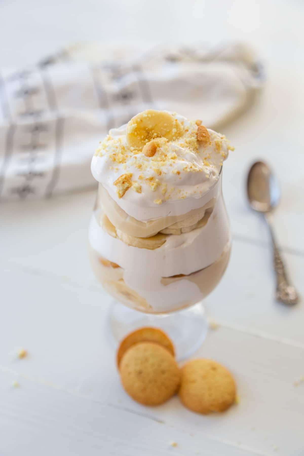 A glass with banana pudding with a slice of banana and crumbled vanilla wafers and a spoon and wafers next to it.