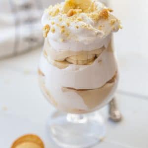 A glass with banana pudding that has layers of the pudding, whipped cream, banana slices, and vanilla wafers.